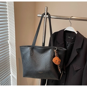 Detail of black tote bag with textured leather, shoulder straps, and a brown teddy bear charm