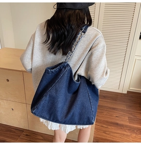 Close-up of a large navy denim tote bag with chain strap worn by a model in a room