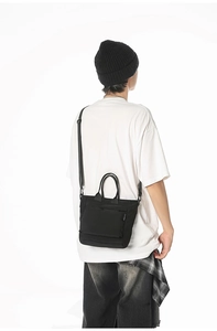Model wearing a black canvas tote bag with adjustable shoulder strap and front pocket