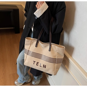 Detail of a large beige canvas tote bag featuring brown stripes and a leather handle