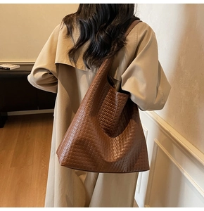 Brown woven leather tote bag shown on shoulder with long beige trench coat in soft light