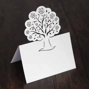 Laser-cut white wedding place card with intricate rose tree design on a wooden background