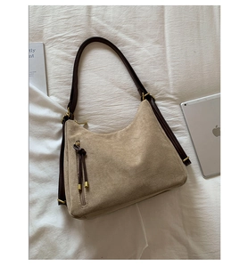 Beige suede hobo bag with brown strap and fringe detail next to tablet and book