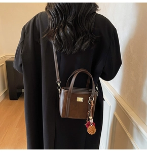 Detail of a dark brown suede crossbody bucket bag with gold metal plate and pompom charm