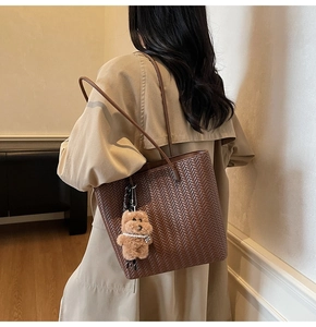 Brown woven tote bag with plush bear charm worn by a model in a cozy interior setting