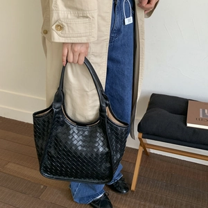 Detail of a large black woven leather tote bag held by a model wearing a trench coat