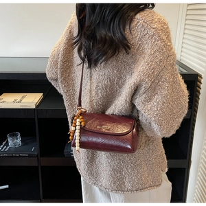 Detail of burgundy leather crossbody bag with wooden bead charm and metal logo on beige knit