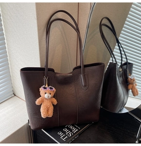 Detail of a dark brown faux leather tote bag with a plush teddy bear charm in soft natural light