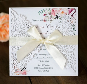 Detail of laser-cut floral lace wedding invitation with white ribbon bow on wood surface