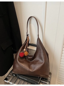 Deep brown large capacity leather tote bag with colorful pompom charm on a silver suitcase