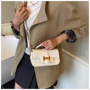 Close-up of cream crossbody bag with gold H-buckle worn with a trench coat