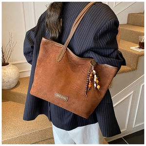 Detail of a brown suede tote bag featuring a beaded tassel and metal logo on the side