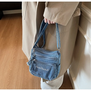 Detail of blue denim crossbody bag with three zipper pockets and embroidered text