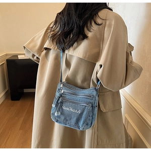 Side view of a blue denim crossbody bag with multiple zippers worn with a tan trench coat