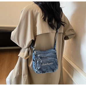 Detail view of a vintage blue denim crossbody bag with multiple zippers worn over a beige coat