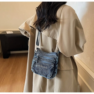 Detail of blue washed denim crossbody bag with multiple zippers and white embroidery