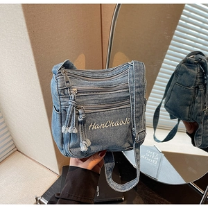 Detail of blue denim crossbody bag with embroidered text and multi-zipper layers