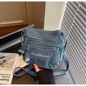 Detail of blue denim crossbody bag featuring multi-zipper layers and white embroidery