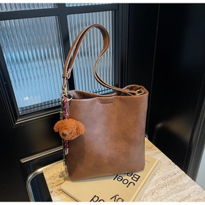 Brown leather bucket bag detail with plush bear charm and contrast rope on stone surface