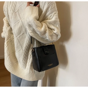 Détail d'un sac seau en cuir noir avec lettrage doré porté sur un gros pull blanc