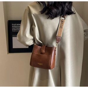 Détail du sac seau en cuir marron avec une large bandoulière brodée et texte classique