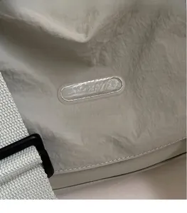 Detail of greyish-white crossbody bag fabric featuring an oval brand logo and strap