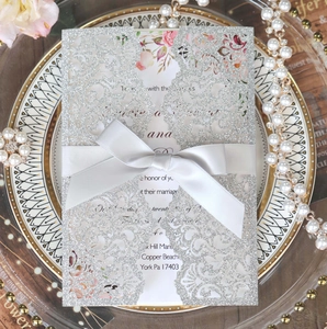 Detail of silver glitter laser cut wedding invitation with white satin bow on a plate