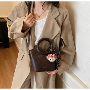 Détail du sac seau marron foncé avec poignée dorée et breloque peluche sur manteau beige