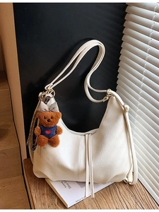 Minimalist beige hobo bag with teddy bear pendant and adjustable straps on a wooden stand