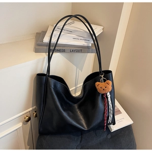 Detail of a black leather tote bag with a brown plush bear charm and long shoulder strap