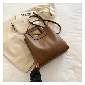 Close-up of brown leather tote bag with unique hollow square handle on a beige blazer