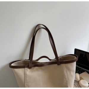 Detail of large canvas tote bag with brown leather trim next to a laptop and headphones