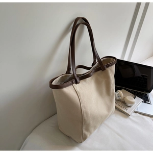 Large beige canvas tote bag with brown leather trim near laptop and art book