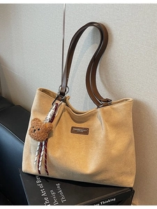 Khaki suede tote bag with brown leather straps and plush bear charm on a dark sofa