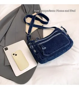 Size comparison of denim crossbody bag with multiple zippers against a phone and tablet
