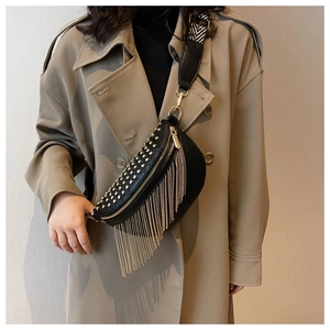 Détail du sac banane noir clouté avec pompons dorés porté sur un trench-coat kaki