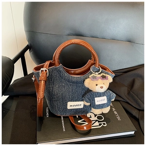 Dettaglio borsa a secchiello in denim blu con manici in pelle marrone e ciondolo orsetto