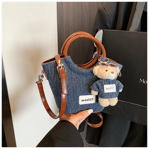 Dettaglio borsa a secchiello in denim con manici in pelle marrone e ciondolo a orsetto
