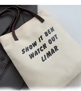 Détail du sac fourre-tout en toile montrant le tissu beige, anses brunes et texte noir