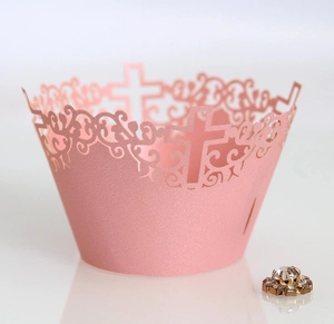 Intricate laser-cut cross and vine patterns on pale pink cupcake wrappers for weddings
