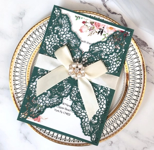 Detail of laser-cut green invitation with satin ribbon and pearl brooch on ceramic plate