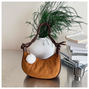 Détail du sac demi-lune en daim fauve avec poignée tressée et pompon blanc sur un bureau