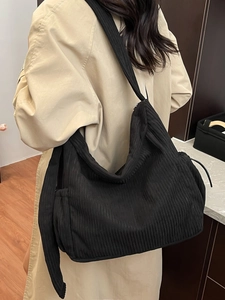 Large black corduroy tote bag with wide strap worn by a model in a long beige trench coat