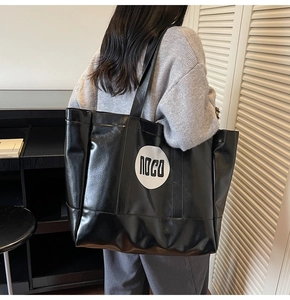 Detail of a large black PU leather tote bag with white logo worn by a model in a room