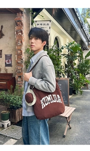 Brown canvas tote bag with white lettering and a mini hat charm in a street setting