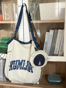Large cream nylon tote bag with navy straps and mini hat pendant on a bookshelf