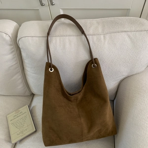 Detail of a brown suede hobo bag with silver eyelets on a white sofa next to a book