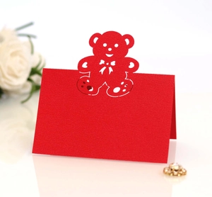 Laser-cut red teddy bear silhouette place card on white glossy surface with white roses