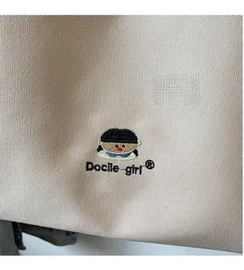 Detail of embroidered cartoon girl character and 'Docile girl' text on beige canvas tote bag