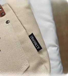 Detail of beige canvas tote with brand label, leather strap, and metal rivets